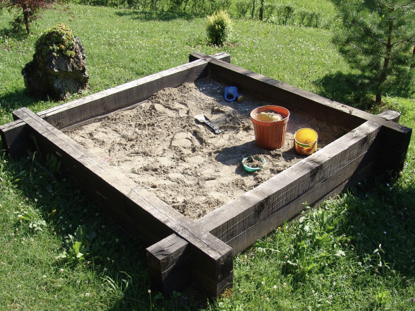 A wooden backyard sandbox full of play sand, with a shovel and a couple of buckets sitting in one corner