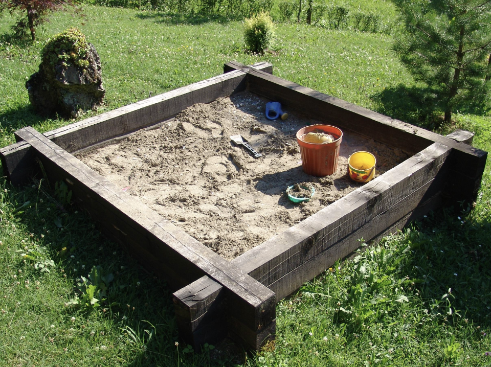 A wooden backyard sandbox full of play sand, with a shovel and a couple of buckets sitting in one corner