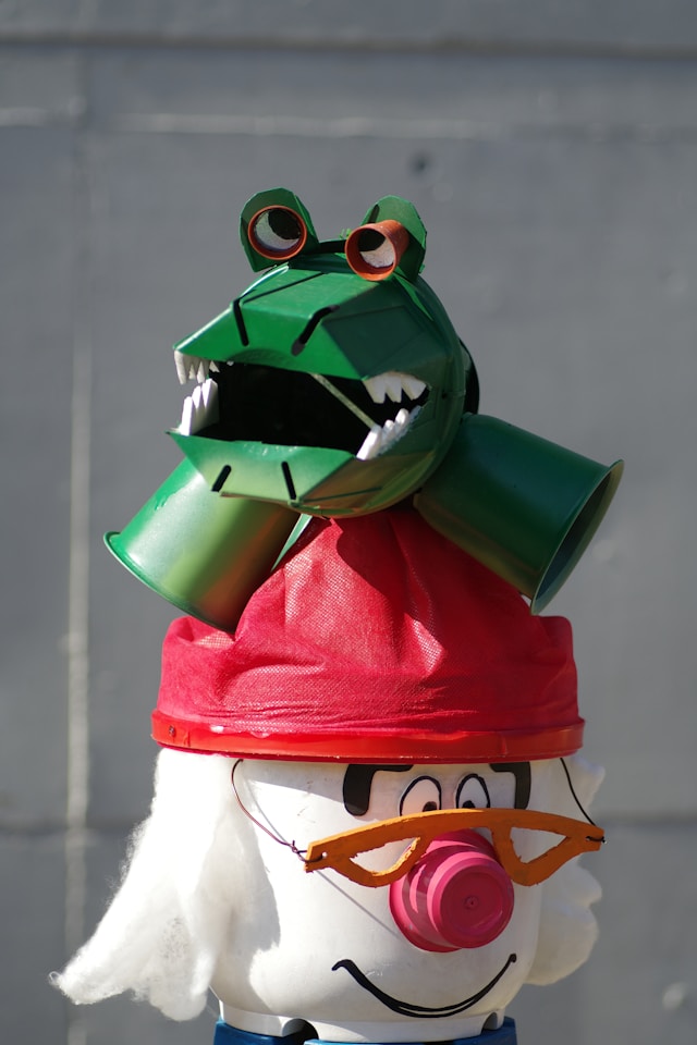 A funky crocodile made out of cups and other materials. It is sitting on top of some other cartoon character, wearing a read hat